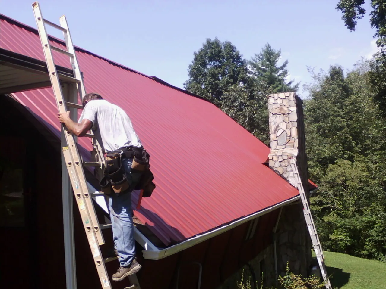 Roofer on a ladder beside a red metal roof and stone chimney — Soffit & Fascia Repair by Roofing Brothers in Rock Stream
